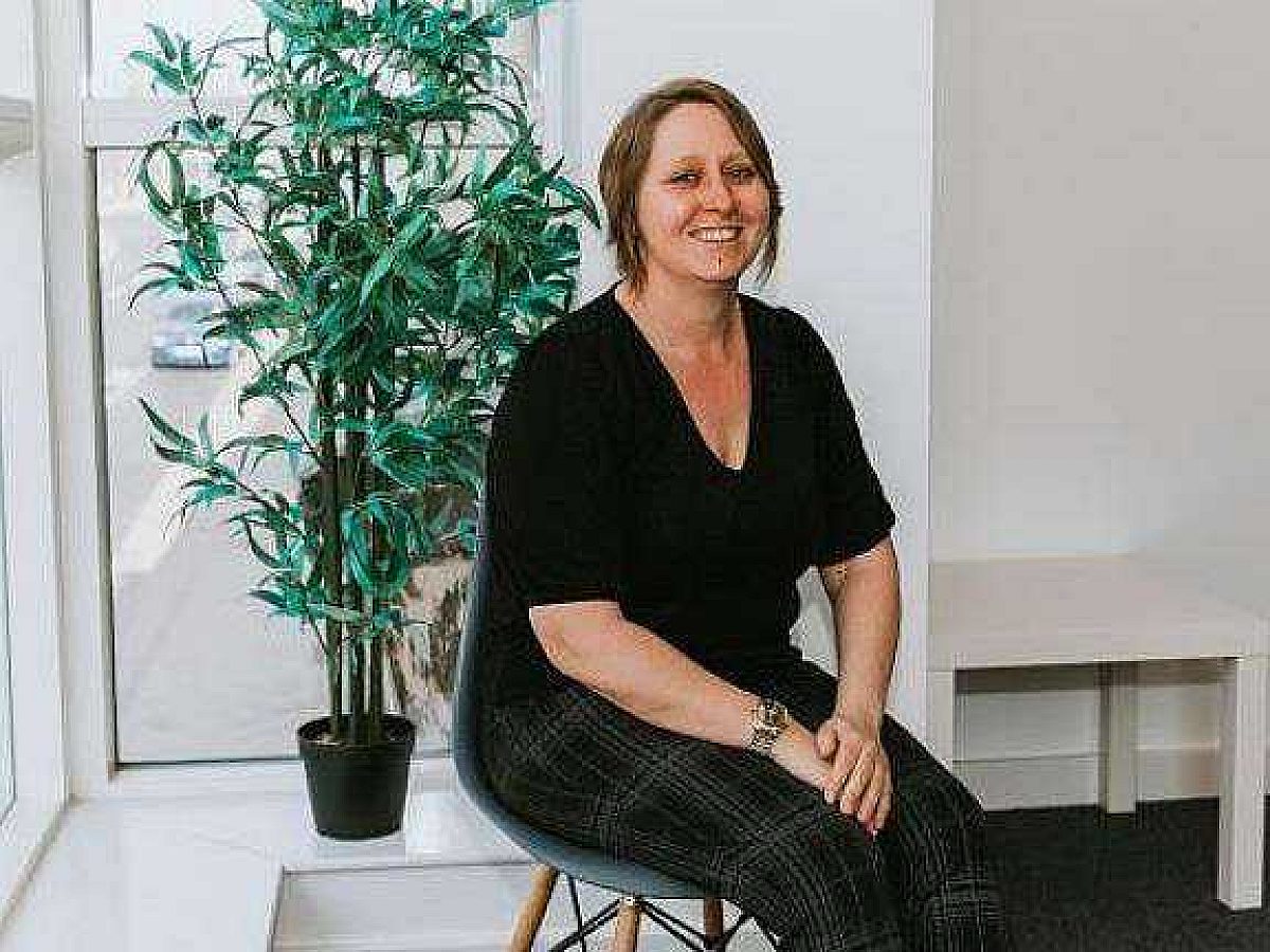 A brown haired woman sits on a chair wearing a black top and smiles at the camera with her hands crossed